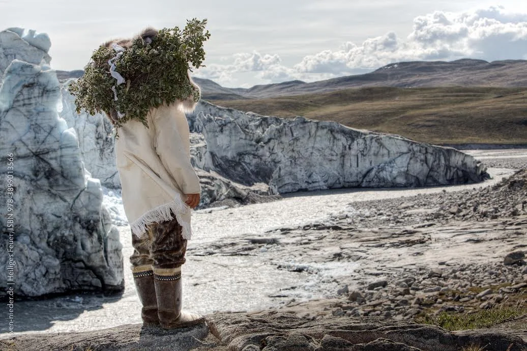 A person in traditional Greenlandic dress with a leafy headdress standing before the glacier and its meltwater river in Kalaallit Nunaat